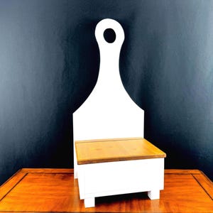 May include: A white wooden bench with a handle-shaped back and a natural wood seat. The bench is painted white with a rectangular seat and four short legs. The handle has a circular opening at the top.