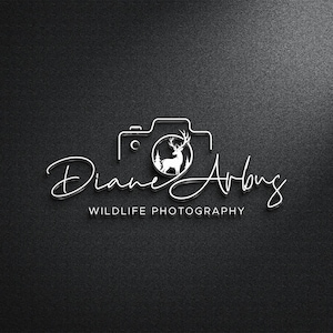 Può includere: Logo bianco su sfondo nero. Presenta un contorno di fotocamera con un cervo all'interno di un cerchio. Il nome "Diane Arbus" è scritto in corsivo sotto la fotocamera, con "WILDLIFE PHOTOGRAPHY" sotto.