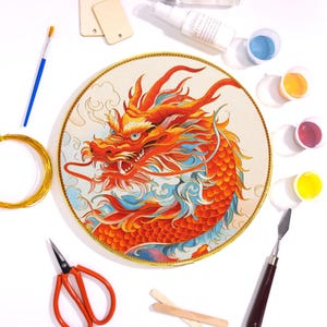 May include: A circular embroidery hoop featuring a vibrant red and orange dragon design with blue accents. The image includes art supplies like paint, brushes, and wire, suggesting a craft project. The dragon is the focal point, with a gold border.