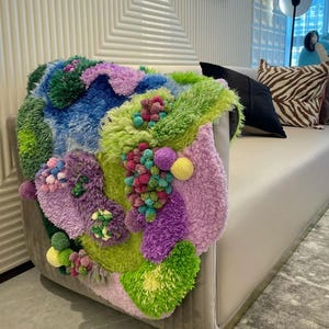 Peut inclure: Une &oelig;uvre d'art textile color&eacute;e et textur&eacute;e drap&eacute;e sur le bras d'un canap&eacute; clair. L'&oelig;uvre pr&eacute;sente un m&eacute;lange vibrant de vert, bleu, violet et rose, avec une vari&eacute;t&eacute; de textures et de formes. Le canap&eacute; est de couleur neutre, dans un salon moderne.
