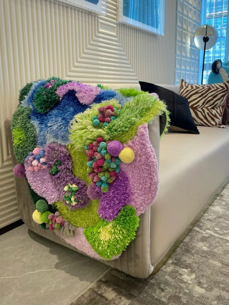 Peut inclure: Une &oelig;uvre d'art textile color&eacute;e et textur&eacute;e drap&eacute;e sur le bras d'un canap&eacute; clair. L'&oelig;uvre pr&eacute;sente un m&eacute;lange vibrant de vert, bleu, violet et rose, avec une vari&eacute;t&eacute; de textures et de formes. Le canap&eacute; est de couleur neutre, dans un salon moderne.