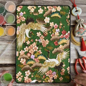 May include: Green tablet case with a detailed design of two cranes in flight amidst cherry blossoms and clouds. The case has a black border. The design incorporates gold, white, pink, and brown colours. Craft supplies and paint containers surround the case.