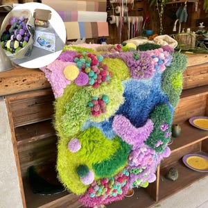 May include: A vibrant, textured textile artwork in shades of green, blue, purple, and pink. The piece features a 3D effect with pom-poms and fluffy textures. A canvas bag with yarn and a small box are visible in the background.
