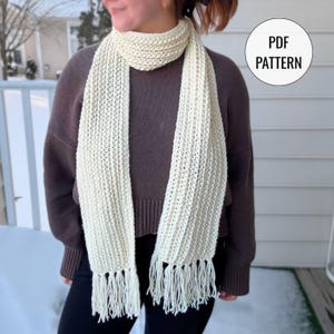May include: Cream-colored, knitted scarf with fringe. The scarf is wrapped around the neck and hangs down the front. The text "PDF PATTERN" is in a white circle in the upper right corner.