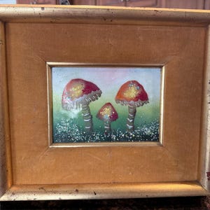 May include: A framed painting of three colorful mushrooms. The mushrooms have red and gold caps with white stems, set against a green and blue background. The painting is in a gold-colored frame.
