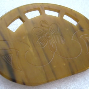 Puede incluir: Un peine para el cabello vintage en tonos marrones y beige, con un diseño floral y cortes decorativos. El peine tiene forma ovalada y está hecho de un material que se asemeja al carey.