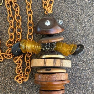 May include: A unique handmade necklace featuring a pendant made of various wooden and amber-colored elements. The pendant is suspended from a gold-colored chain. The pendant has a layered design with a variety of shapes and textures.