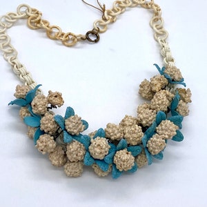 Pode incluir: Um colar vintage com corrente creme e arranjo floral. Apresenta aglomerados de contas bege e detalhes de flores azul claro. O fecho é bronze. Uma peça de joalharia única.