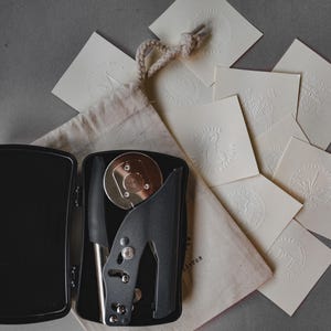 May include: A black embossing seal kit with a silver metal die and handle, open inside a black tin. Several cream-colored embossed cards with various designs are scattered around the kit, along with a small canvas bag.
