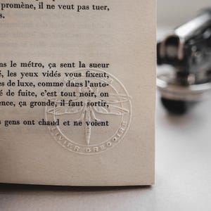 May include: Close-up of a book page with French text. A circular embossed design of a dragonfly is visible. The page is off-white, and the text is in black. A blurred, metallic object is in the background.