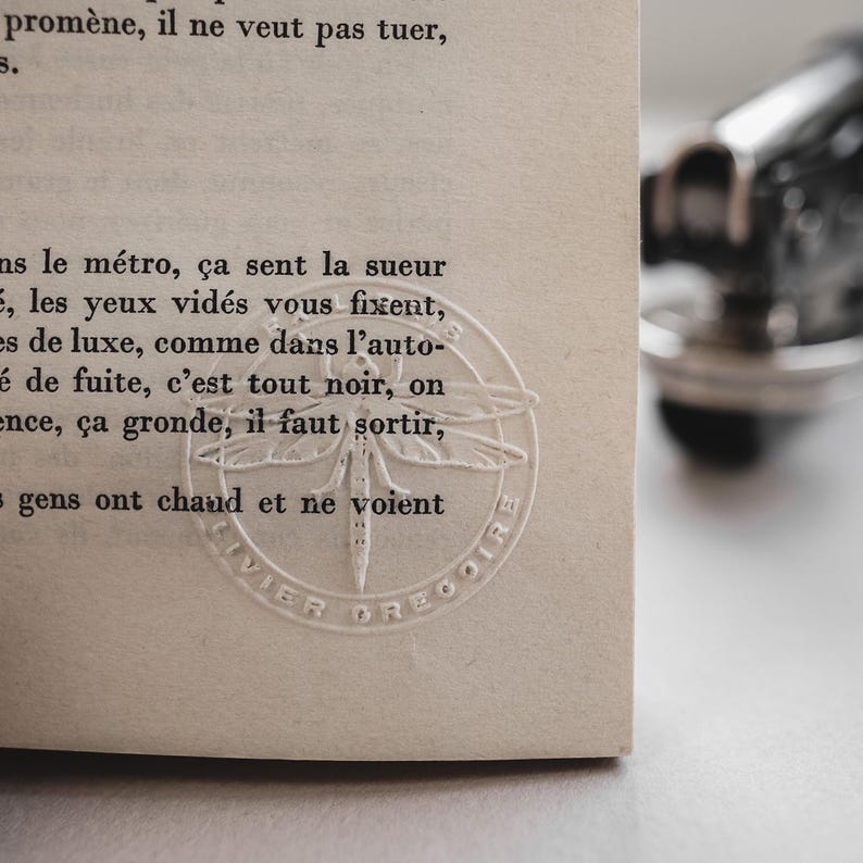 May include: Close-up of a book page with French text. A circular embossed design of a dragonfly is visible. The page is off-white, and the text is in black. A blurred, metallic object is in the background.