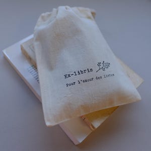 May include: A natural canvas drawstring bag with the text "Ex-libris Pour l'amour des livres" printed in black. The bag rests on top of two books with white and beige covers.