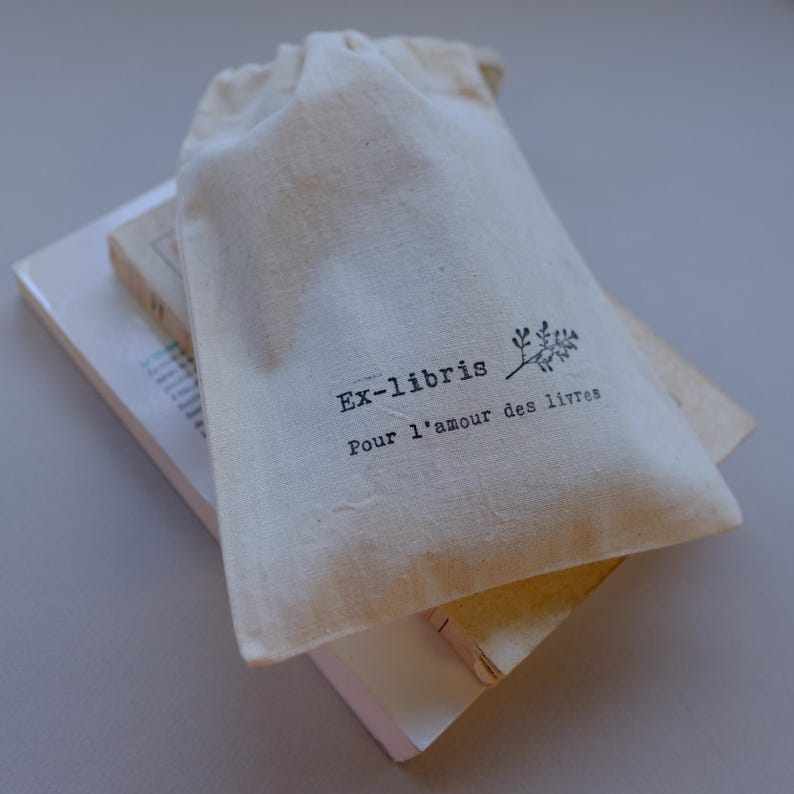 May include: A natural canvas drawstring bag with the text "Ex-libris Pour l'amour des livres" printed in black. The bag rests on top of two books with white and beige covers.