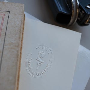May include: A close-up of a book and a white card with an embossed design. The card features the text "Ex-libris" above a rose illustration, with "Caroline Grisham" below. A blue book press is in the background.