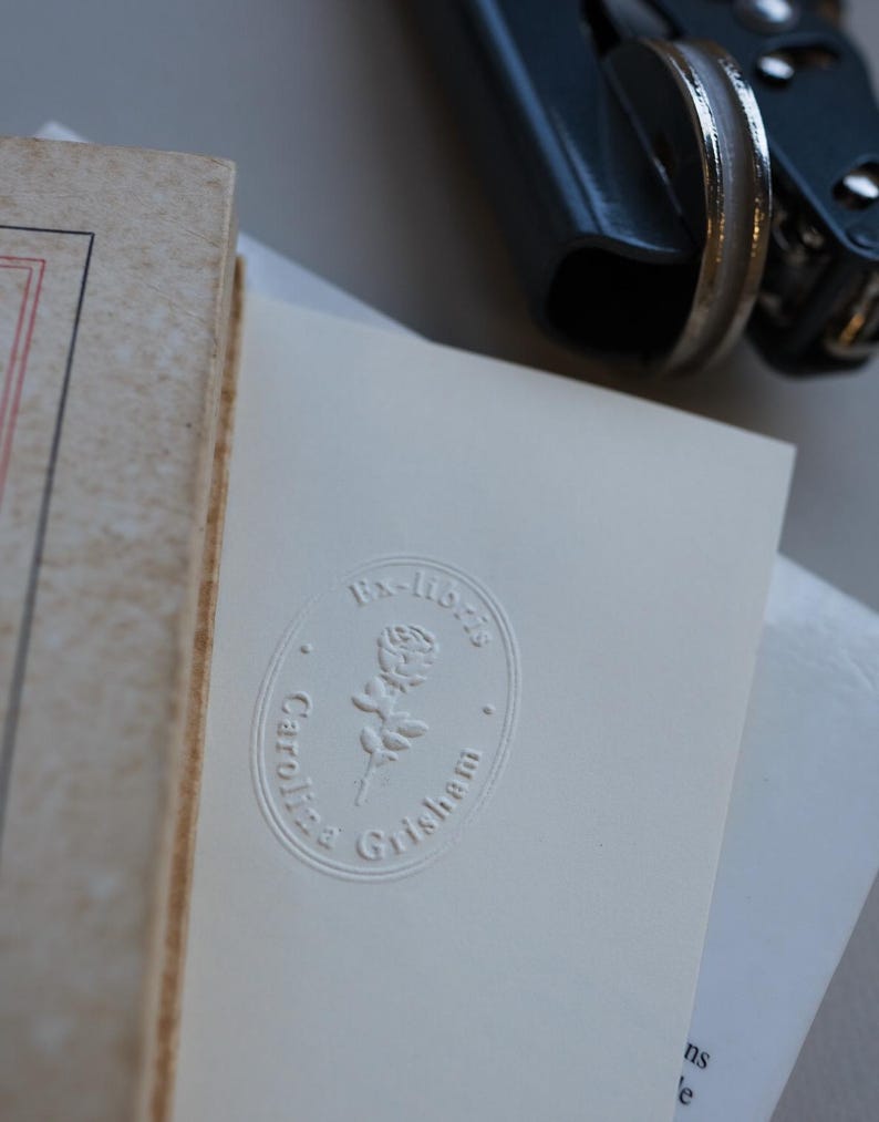May include: A close-up of a book and a white card with an embossed design. The card features the text "Ex-libris" above a rose illustration, with "Caroline Grisham" below. A blue book press is in the background.