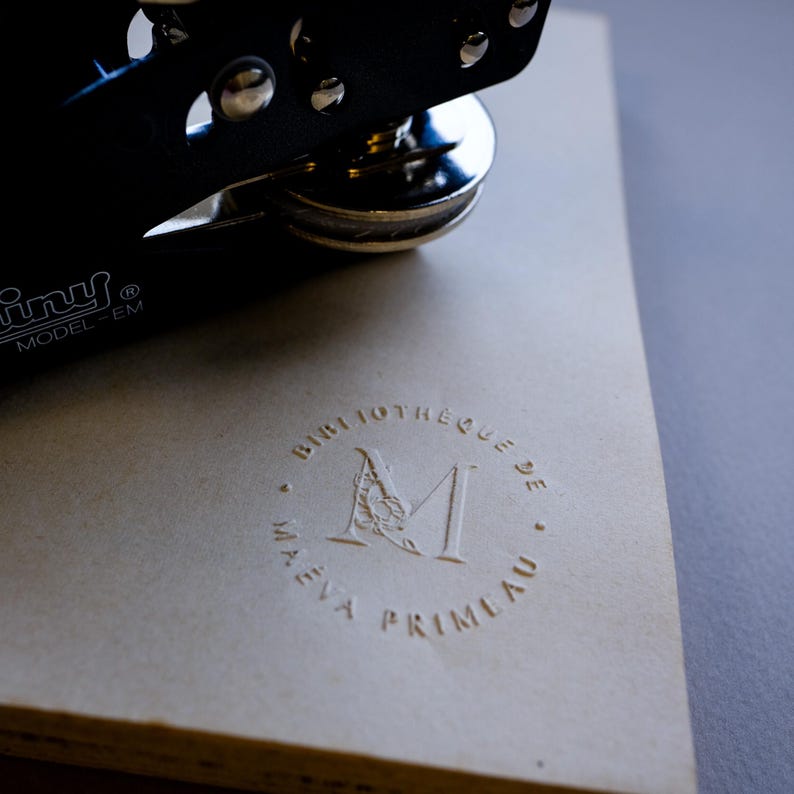 May include: A black Tiny Model-EM embosser rests on a beige paper surface. The paper is embossed with a circular design that includes the text "BIBLIOTHEQUE DE M. EVA PRIMEAU". The embosser is partially visible, with its silver metal components.