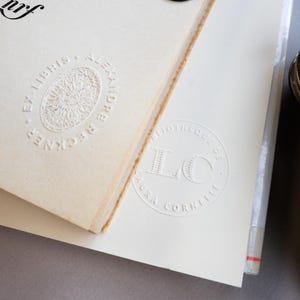 May include: Close-up of two cream-colored book pages with embossed text and designs. One page features "EX LIBRIS ALEXANDRE RECKNER" around a central sunburst design. The other page has "BIBLIOTHEQUE DE LAURA CORNELII" with "LC" in the center.