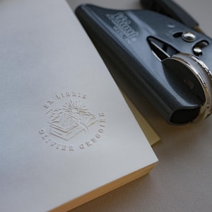 May include: A white book with embossed text and a gray stapler. The book has the words "EX LIBRIS OLIVIER GREGOIRE" and a sun and mountain design. The stapler is gray and has the word "Swingline" on it.