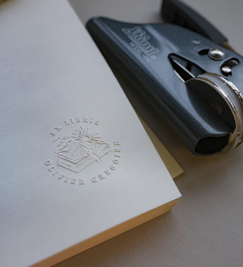 May include: A white book with embossed text and a gray stapler. The book has the words "EX LIBRIS OLIVIER GREGOIRE" and a sun and mountain design. The stapler is gray and has the word "Swingline" on it.