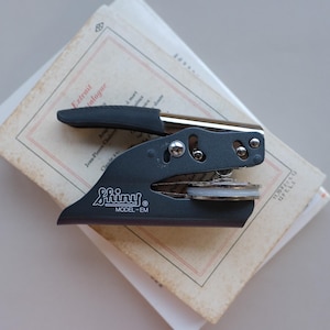 May include: A black Shiny Model-EM embosser sits atop a stack of aged paper. The embosser has a silver handle and a black grip. The paper has red and black text. The embosser is a stationery item.