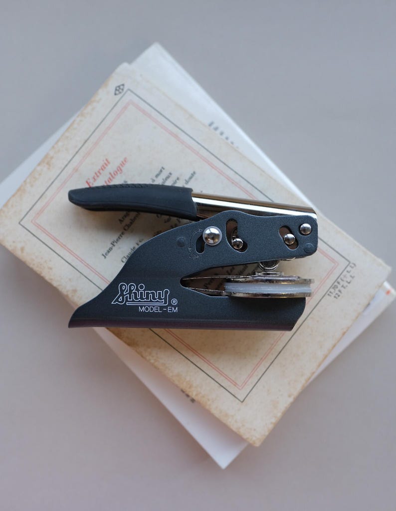 May include: A black Shiny Model-EM embosser sits atop a stack of aged paper. The embosser has a silver handle and a black grip. The paper has red and black text. The embosser is a stationery item.