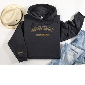Puede incluir: Sudadera con capucha gris oscuro con el texto dorado "GENERATION X" y "ZERO PATIENCE MODE". La manga tiene la palabra "SERIOUSLY" bordada en dorado. Un sombrero de paja y unos vaqueros también están en la imagen.