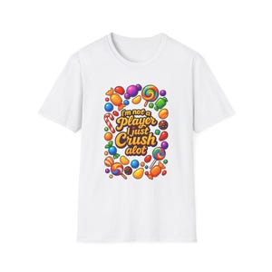 Puede incluir: Camiseta blanca con un diseño colorido de caramelos y el texto "I'm not a player I just crush a lot." El diseño incluye piruletas, caramelos y otros dulces de varios colores.