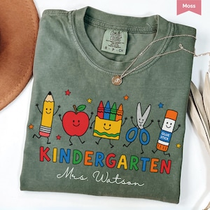 May include: A moss green short-sleeve t-shirt featuring a graphic of a smiling pencil, apple, crayon box, scissors, and glue stick. The text reads KINDERGARTEN and Mrs. Watson. The shirt has a crew neckline and a Comfort Colors brand label.