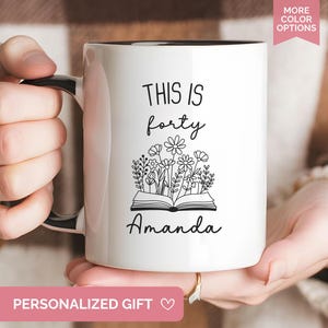 May include: White ceramic mug with a black interior and handle. The mug features the text "THIS IS forty" above a floral illustration and the name "Amanda". The mug is held by a person.