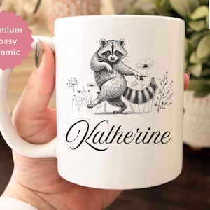 May include: White ceramic mug with a black and white illustration of a raccoon standing among flowers. The mug has the name "Katherine" in a cursive font. The mug is labeled "11 oz All White" and "Premium glossy ceramic".