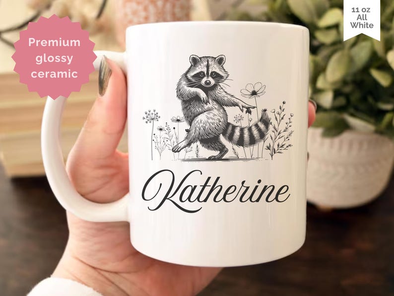 May include: White ceramic mug with a black and white illustration of a raccoon standing among flowers. The mug has the name "Katherine" in a cursive font. The mug is labeled "11 oz All White" and "Premium glossy ceramic".
