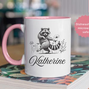May include: A white ceramic mug with a pink handle and interior, featuring a black and white illustration of a dancing raccoon surrounded by wildflowers. The name Katherine is printed in black script below the raccoon. The mug holds 11 oz of liquid.