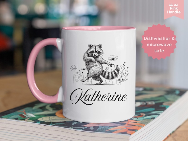 May include: A white ceramic mug with a pink handle and interior, featuring a black and white illustration of a dancing raccoon surrounded by wildflowers. The name Katherine is printed in black script below the raccoon. The mug holds 11 oz of liquid.