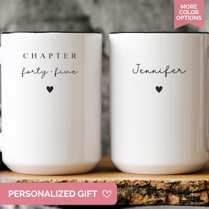 May include: Two white ceramic mugs with black handles and rims. One mug reads "CHAPTER forty-five" with a heart symbol, the other reads "Jennifer" with a heart. The mugs are on a wooden surface.