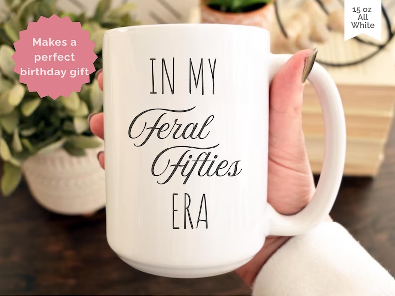 May include: White ceramic mug with the text "IN MY Feral Fifties ERA" in black script. The mug is held by a person. A pink sticker with the text "Makes a perfect birthday gift" is in the background. The mug is 15 oz.