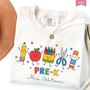 May include: An ivory t-shirt featuring a graphic of a smiling pencil, apple, crayon box, scissors, and glue stick. Below the characters, the text reads PRE-K and Mrs. Watson in a script font. The shirt has a Comfort Colors brand neck label.