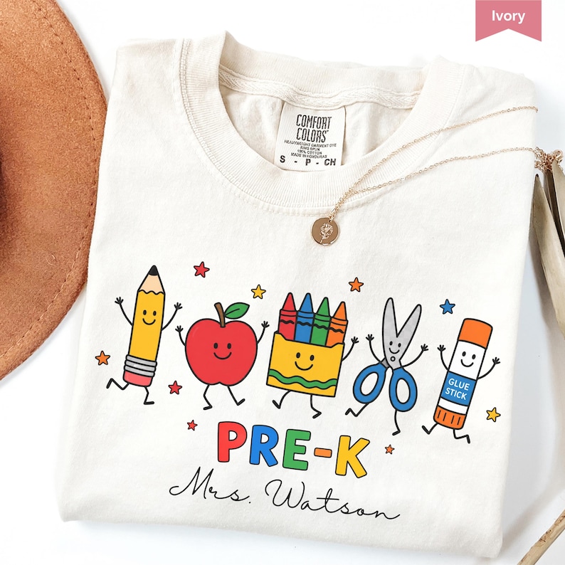 May include: An ivory t-shirt featuring a graphic of a smiling pencil, apple, crayon box, scissors, and glue stick. Below the characters, the text reads PRE-K and Mrs. Watson in a script font. The shirt has a Comfort Colors brand neck label.
