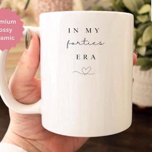 May include: White ceramic mug with the text "IN MY forties ERA" in black script, and a small heart. The mug is 11 oz and has a glossy finish. The mug is held by a person.