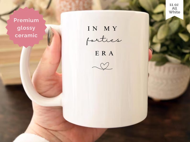 May include: White ceramic mug with the text "IN MY forties ERA" in black script, and a small heart. The mug is 11 oz and has a glossy finish. The mug is held by a person.