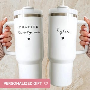 May include: Two white insulated tumblers with handles, each held by a person. One tumbler reads "CHAPTER twenty-one" with a heart, the other says "Taylor" with a heart. Both have white lids and silver trim. The tumblers are personalized gifts.