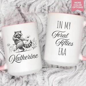 May include: Two white ceramic mugs with pink handles and rims. One mug features a raccoon illustration and the name "Katherine". The other mug has the text "IN MY Feral Fifties ERA". Each mug holds 15 oz.