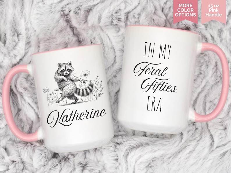 May include: Two white ceramic mugs with pink handles and rims. One mug features a raccoon illustration and the name "Katherine". The other mug has the text "IN MY Feral Fifties ERA". Each mug holds 15 oz.