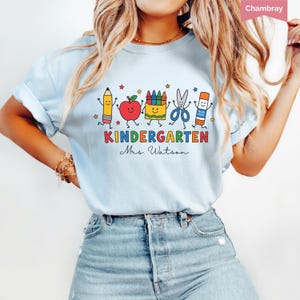 Personalized kindergarten teacher shirt with colorful school supplies, apple, crayons, scissors, glue stick, and custom teacher name, perfect back to school gift.