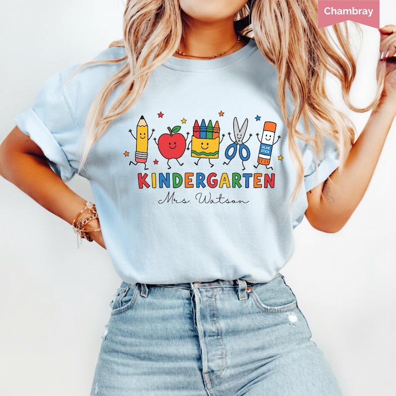 Personalized kindergarten teacher shirt with colorful school supplies, apple, crayons, scissors, glue stick, and custom teacher name, perfect back to school gift.