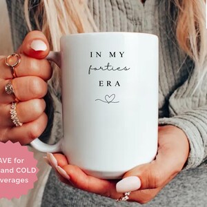 May include: A white ceramic mug with the text "IN MY forties ERA" in black script and a small heart. The mug is held by a person. The mug is labeled "15 oz All White". A pink sticker says "SAVE for HOT and COLD beverages".