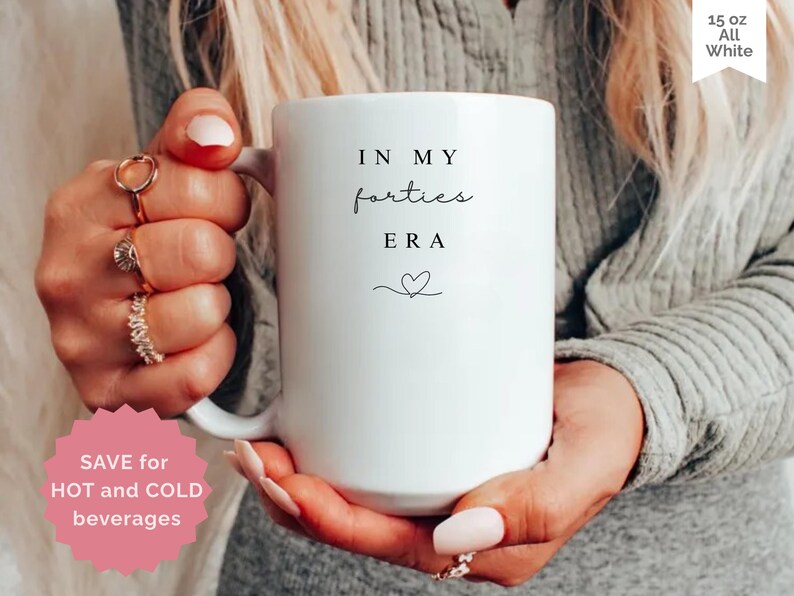 May include: A white ceramic mug with the text "IN MY forties ERA" in black script and a small heart. The mug is held by a person. The mug is labeled "15 oz All White". A pink sticker says "SAVE for HOT and COLD beverages".