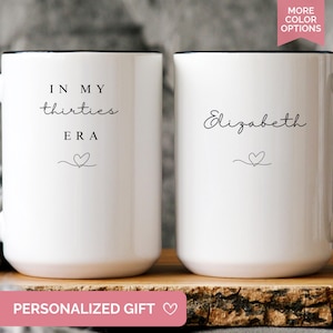 May include: Two white ceramic mugs with black handles and rims. One mug reads "IN MY thirties ERA" with a heart symbol. The other mug says "Elizabeth" with a heart symbol. The mugs are on a wooden surface.