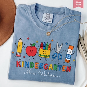 Personalized kindergarten teacher shirt with colorful school supplies, apple, crayons, scissors, glue stick, and custom teacher name, perfect back to school gift.