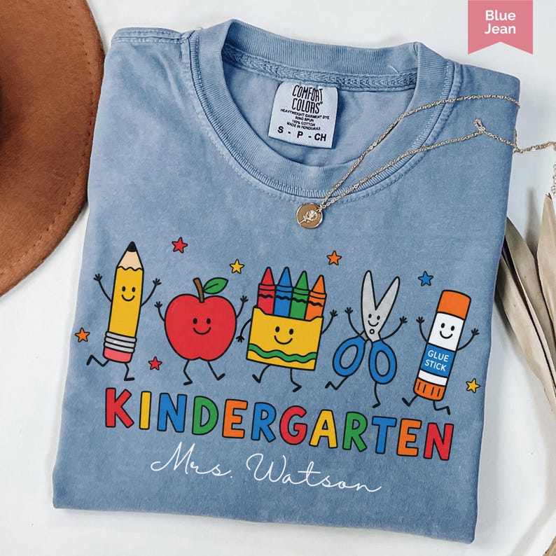 Personalized kindergarten teacher shirt with colorful school supplies, apple, crayons, scissors, glue stick, and custom teacher name, perfect back to school gift.