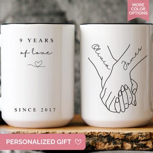 May include: Two white ceramic mugs with black handles and rims. One mug reads "9 years of love" and "Since 2017". The other mug features a line drawing of two hands holding each other with the names "Olivia" and "James".